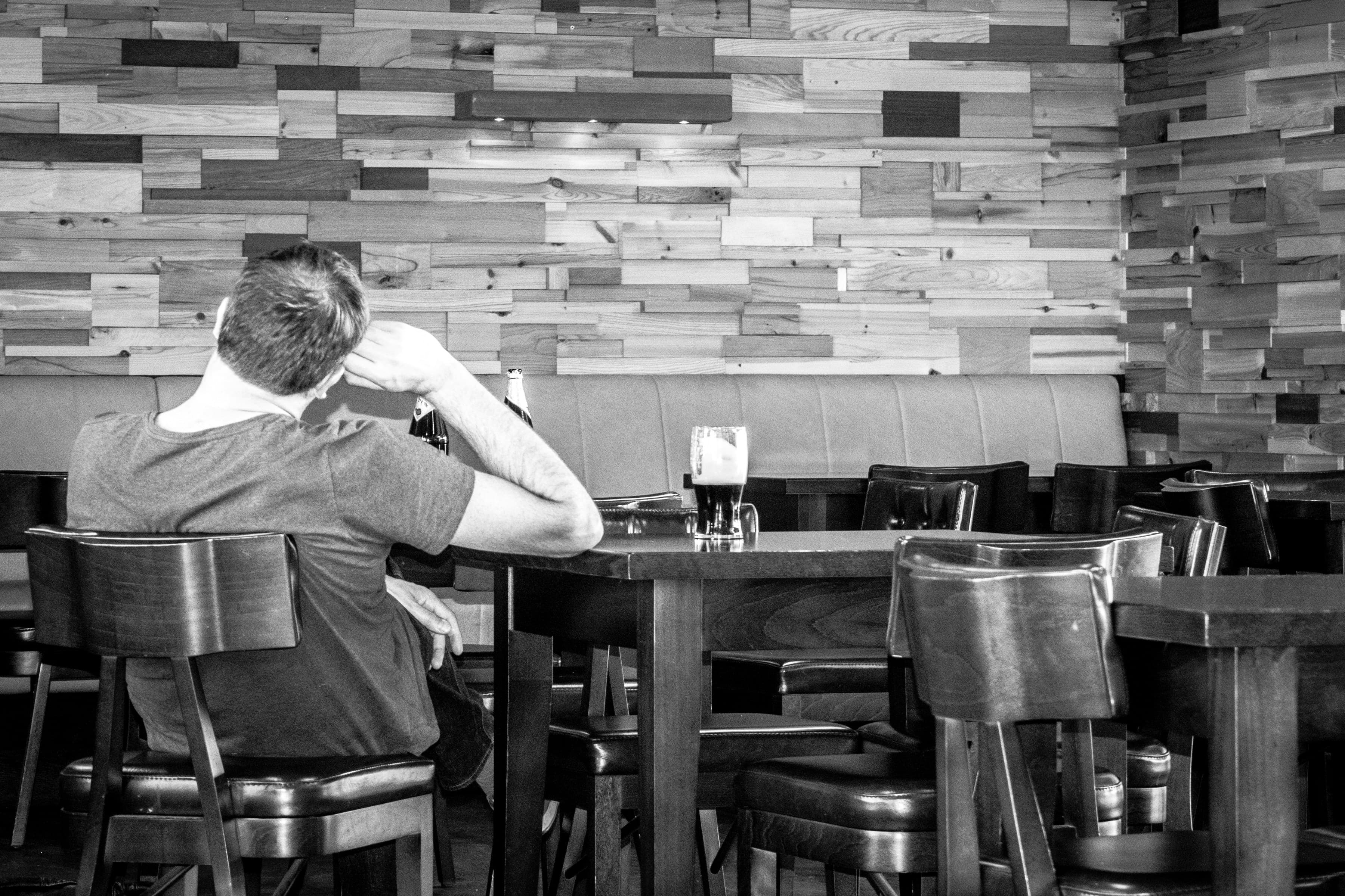 A man sitting alone at a restaurant table, back to camera, with a beer in front of him