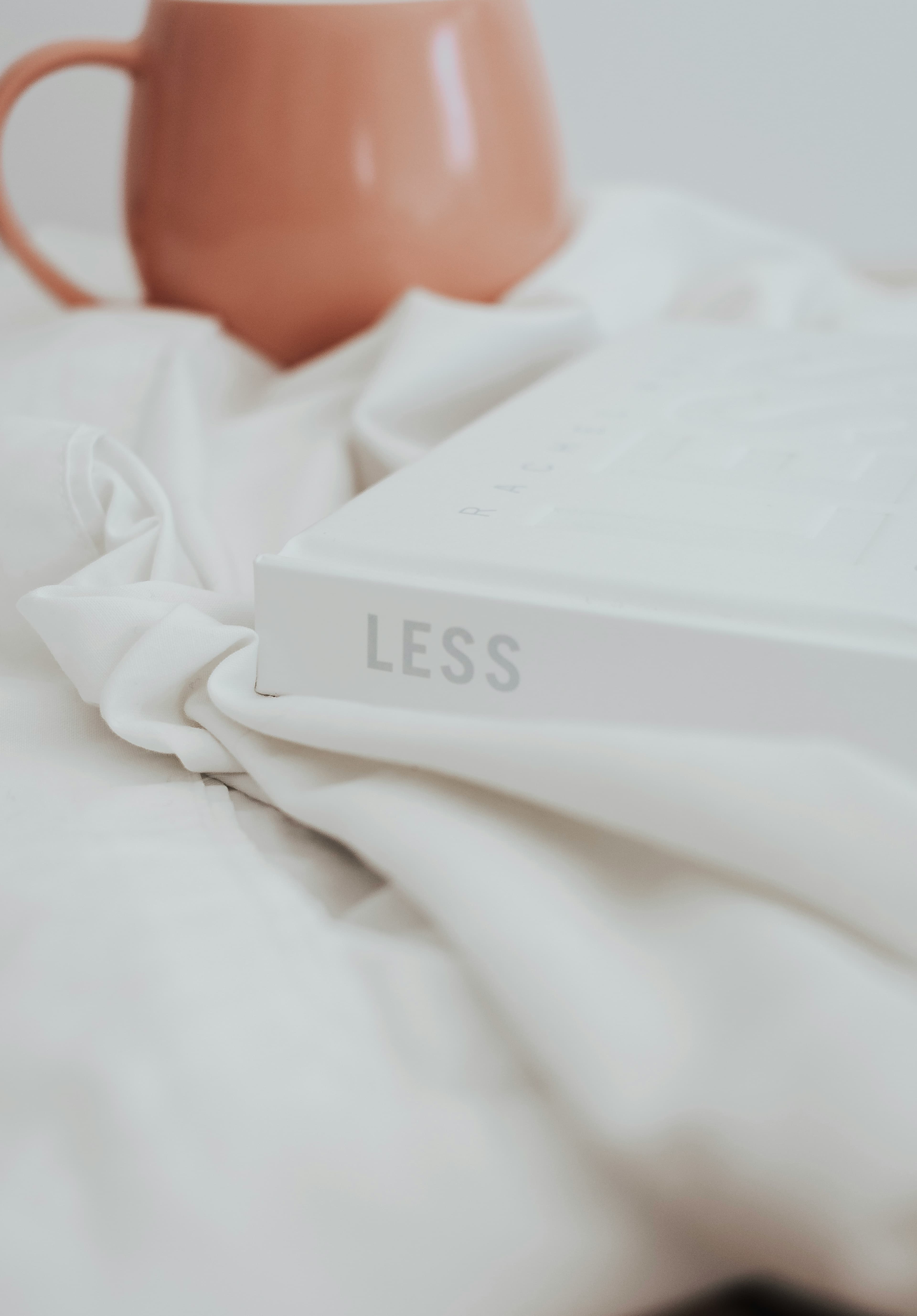 A book titled LESS resting on white fabric next to a pink mug