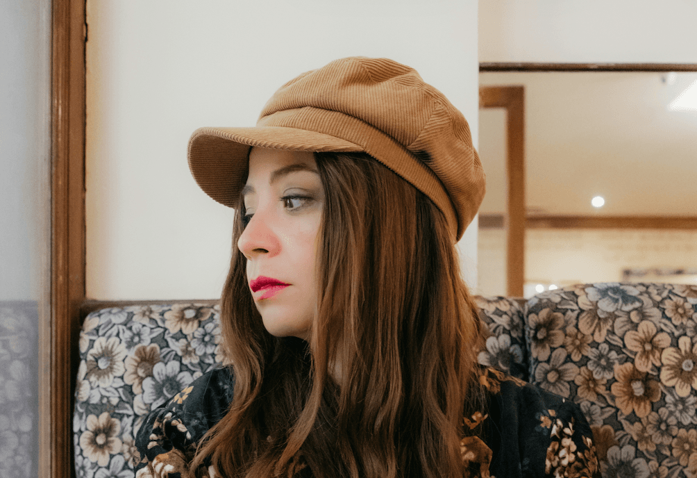A woman in a corduroy cap sits in a floral restaurant booth, holding a cappuccino and looking away in thought